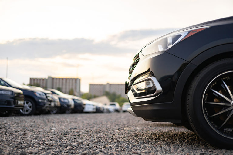A row of cars parked outside on a gravel lot, featuring a close-up of a black vehicle's front.